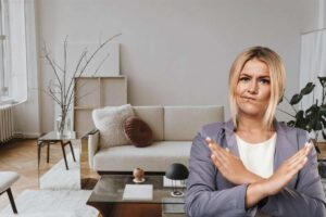 Donna X con le braccia e sul fondo salotto con divano beige e tavolino, piante