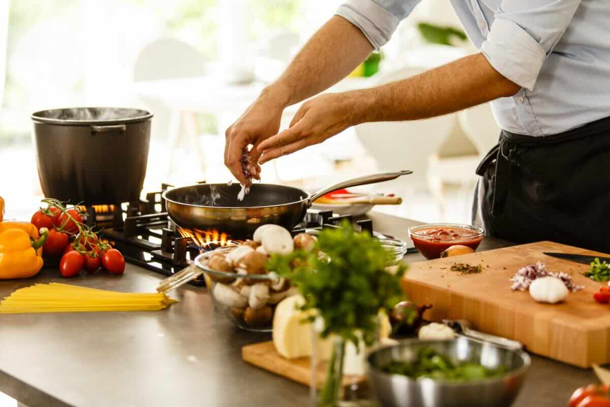 Uomo che versa qualcosa in padella, vari ingredienti sul piano da lavoro